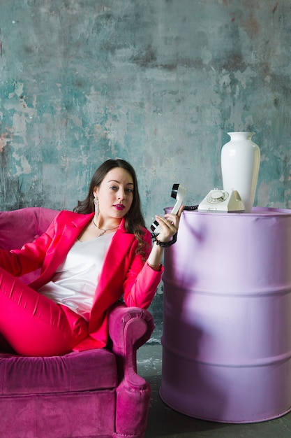 Belle jeune femme en costume rose assis sur une chaise rose avec un vieux téléphone à l'intérieur du loft