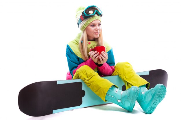 Belle jeune femme en costume de ski et lunettes de ski s'asseoir sur la planche à neige, tenez la tasse rouge