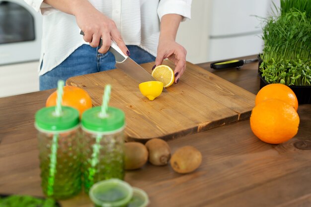 Belle jeune femme coupe un citron dans la cuisine sur la table pour un smoothie vitaminé à base de microgreen et de fruits.