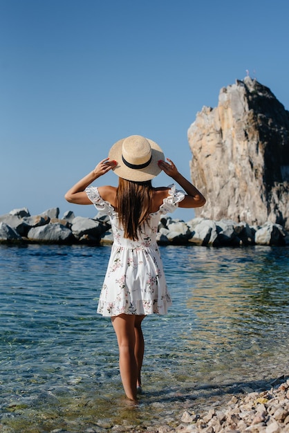 Une belle jeune femme dans un chapeau et une robe légère avec le dos marche le long du rivage de l'océan sur fond d'énormes rochers par une journée ensoleillée. Tourisme et voyages de vacances.