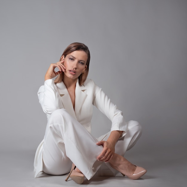 Belle jeune femme dans un costume blanc à la mode et des chaussures à talons hauts beiges, assise sur le sol, un portrait de mode en studio sur fond gris