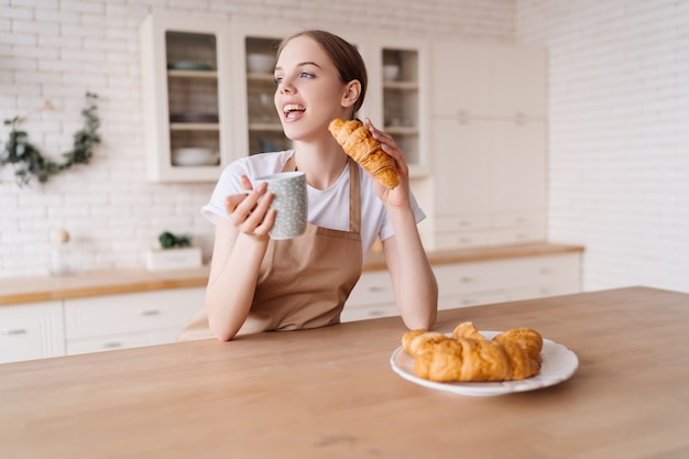 Belle jeune femme dans la cuisine dans un tablier avec café et croissant profite de sa matinée