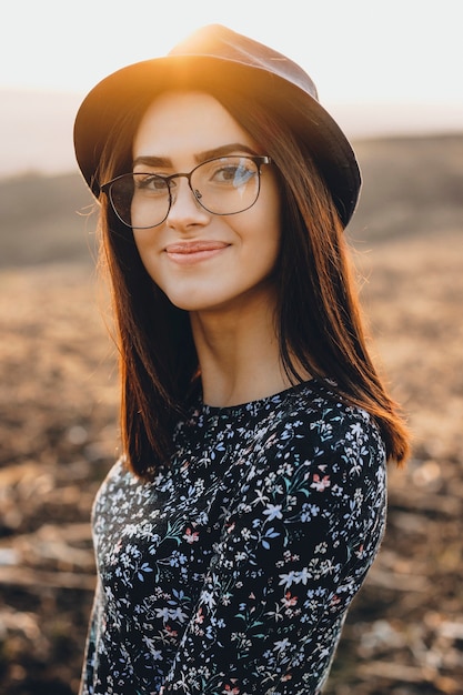 Belle jeune femme, dans, élégant, chapeau, et, lunettes, sourire, et, regarder appareil-photo, quoique, debout, sur, arrière-plan flou, de, champ, pendant, sunset., Amical, femme, dans, soir, campagne