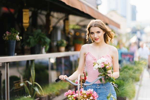 Une belle jeune femme dans une jupe en jean et un haut rose se promène dans les rues de la ville en été avec un vélo