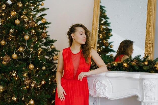 Belle jeune femme dans une longue robe rouge près de la cheminée et de l'arbre de Noël