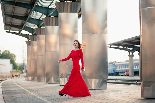 La belle jeune femme dans une longue robe rouge se tient sur la plate-forme de la station près du chemin de fer