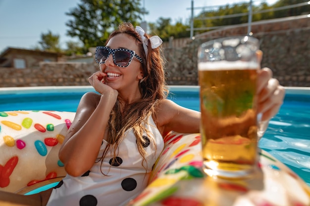 Belle jeune femme dans la piscine nage sur un beignet gonflable et s'amuse avec un verre de bière en vacances.