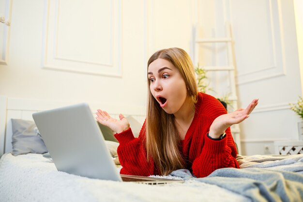 Belle jeune femme dans un pull rouge tapant sur un ordinateur portable assis sur un lit dans une pièce lumineuse. Travailler à distance dans une atmosphère familiale pendant la quarantaine