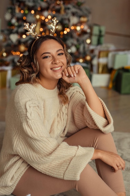 Belle jeune femme dans un pull tricoté volumineux attendant le Nouvel An à la maison à l'intérieur près de l'arbre de Noël