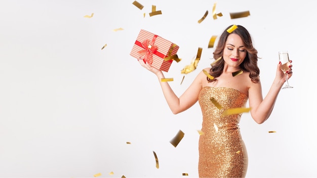 Belle jeune femme dans une robe dorée boit du champagne, tenant une boîte-cadeau rouge, s'amusant lors d'une fête avec des confettis dorés sur fond blanc