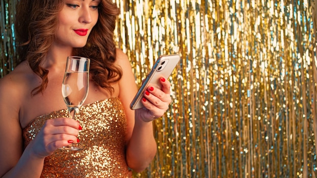 Belle jeune femme dans une robe dorée boit du champagne, tenant un smartphone, s'amusant lors d'une fête sur un fond d'or brillant