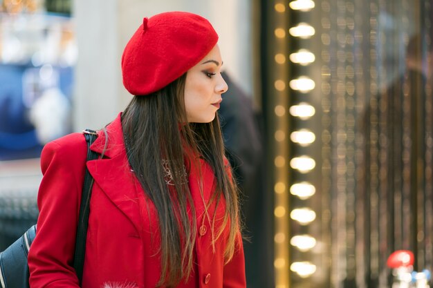 Belle jeune femme dans une robe rouge et un chapeau tenant des sacs à provisions et vérifiant un magasin en plein air dans un cadre de la ville moderne