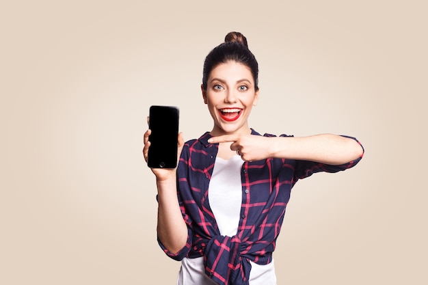 Belle jeune femme dans un style décontracté tenant un téléphone en regardant la caméra et montrant l'écran du téléphone avec le doigt. tourné en studio sur fond beige.