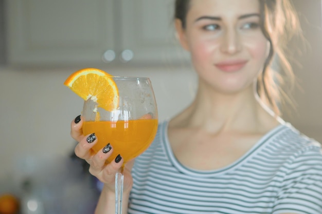 Photo une belle jeune femme dans un t-shirt rayé tient dans sa main un verre avec une tranche d'orange et du jus dans la cuisine