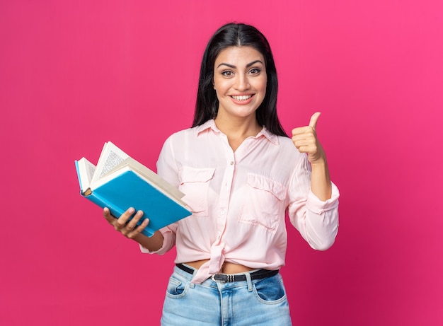 Belle jeune femme dans des vêtements décontractés tenant un livre heureux et positif souriant montrant gaiement les pouces vers le haut debout sur le rose