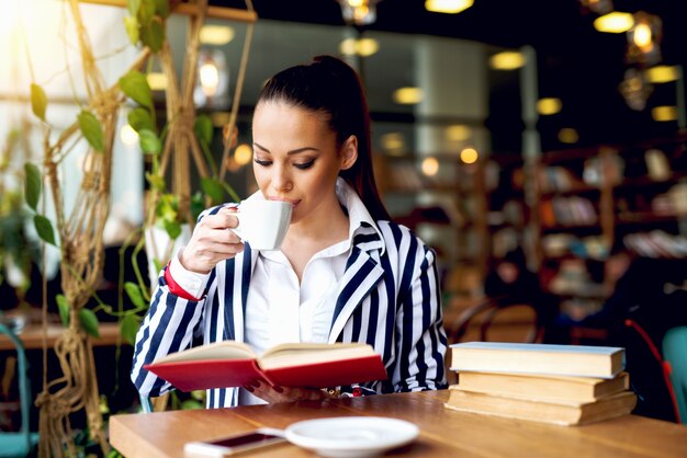 Belle jeune femme dans des vêtements élégants, boire du café pendant la lecture