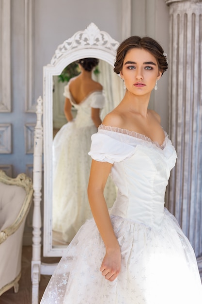 Photo belle jeune femme debout dans une robe blanche près du miroir.