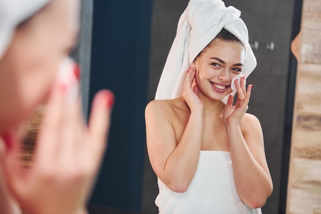 Belle jeune femme debout dans la salle de bain, regardant dans le miroir et prenant soin de son visage.