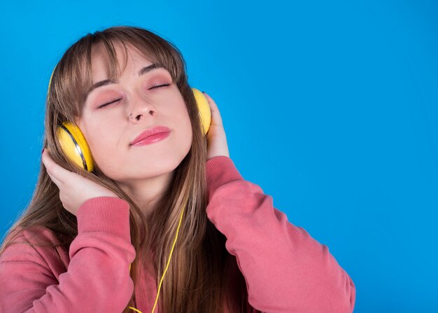 Une belle jeune femme écoute de la musique avec des écouteurs avec les yeux fermés, fond bleu