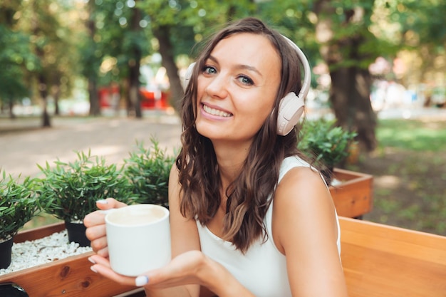 Une belle jeune femme avec des écouteurs est assise à une table dans un café d'été et boit du café ou du thé génération z