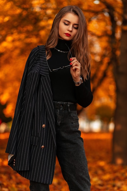 Belle jeune femme élégante dans un costume noir à la mode avec un blazer et un pull dans un parc d'automne à l'extérieur. Style et mode féminins élégants