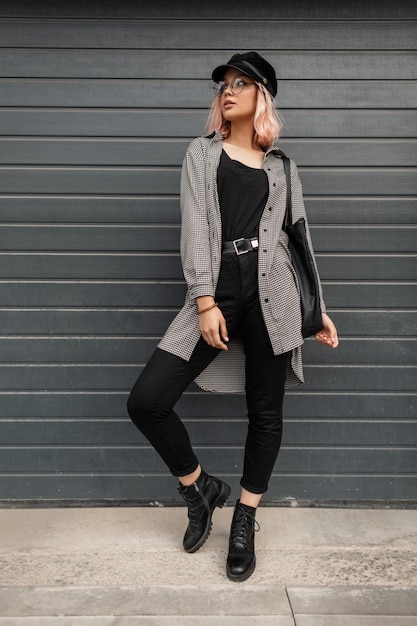 Belle jeune femme élégante avec des lunettes et un chapeau vintage dans des vêtements décontractés à la mode avec une chemise et un jean se tient près de la porte dans la rue