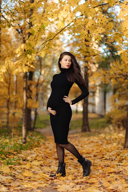 Belle jeune femme enceinte aux cheveux noirs dans une robe moulante noire posant sur une prairie d'automne