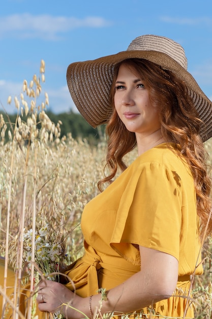 Une belle jeune femme enceinte dans une robe jaune et un chapeau se promène dans un champ de blé orange sur une journée d'été ensoleillée