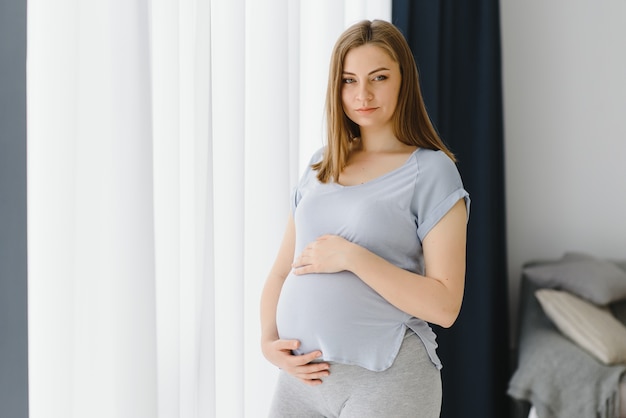 Belle jeune femme enceinte debout à la maison près de la fenêtre.