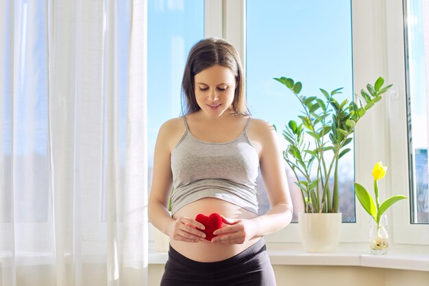 Belle jeune femme enceinte montrant un cœur symbolique, femme posant à la maison près de la fenêtre. Avec amour pour le bébé