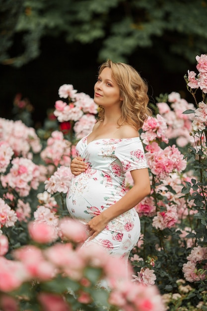 Une belle jeune femme enceinte se promène dans une roseraie. Portrait d'une femme enceinte vêtue d'une robe. Été.
