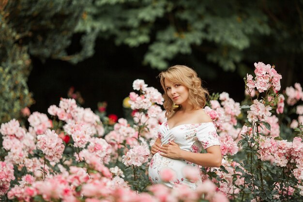 Une belle jeune femme enceinte se promène dans une roseraie. Portrait d'une femme enceinte vêtue d'une robe. Été.