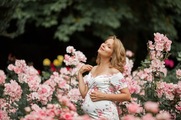 Une belle jeune femme enceinte se promène dans une roseraie. Portrait d'une femme enceinte vêtue d'une robe. Été.