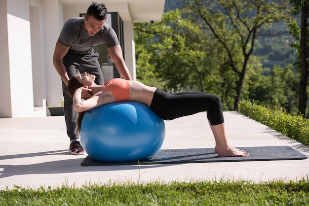 belle jeune femme et entraîneur personnel faisant de l'exercice avec une balle de pilates devant sa maison de luxe