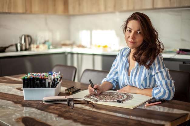 Belle jeune femme est assise dans la cuisine de la maison et dessine un dessin zen mandala à colorier