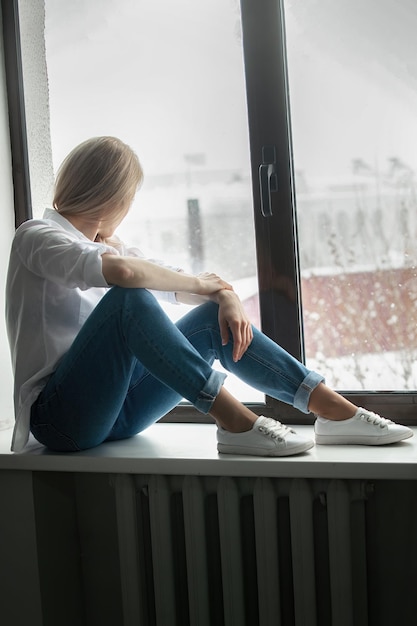 belle jeune femme est assise sur le rebord de la fenêtre dans une chemise blanche et un jean et regarde par la fenêtre