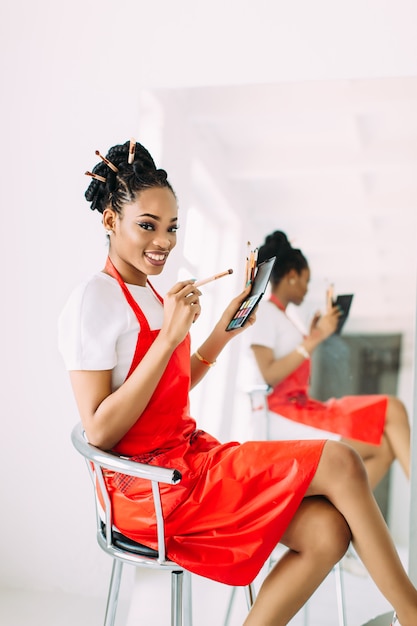 Belle jeune femme esthéticienne afro-américaine tenant ensemble de pinceaux de maquillage et regarder dans le miroir