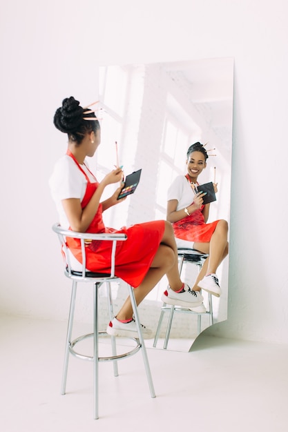 Belle jeune femme esthéticienne afro-américaine tenant ensemble de pinceaux de maquillage et regarder dans le miroir