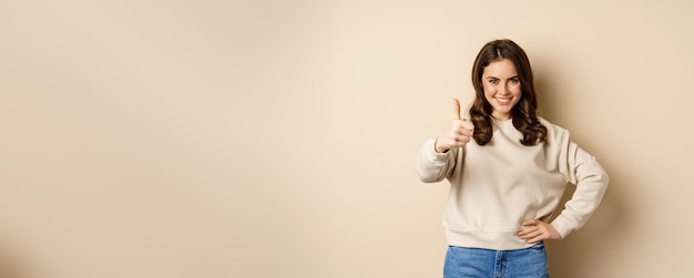 Photo belle jeune femme étudiante montrant les pouces vers le haut en approbation recommandant un magasin debout sur beige b