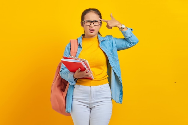 Belle jeune femme étudiante en vêtements en denim avec sac à dos tenant un carnet de notes et Pointez les doigts vers la tête comme si elle se tirait dessus isolée sur fond jaune