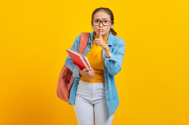 Belle jeune femme étudiante en vêtements en denim avec sac à dos et tenant les doigts du livre couvrant les lèvres faisant un geste silencieux sur fond jaune Éducation au concept de collège universitaire de lycée