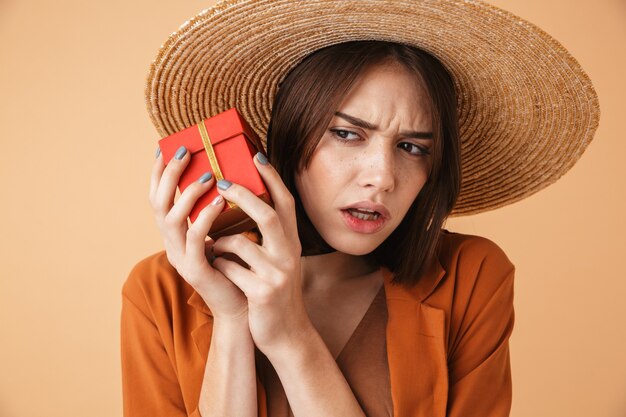 Belle jeune femme excitée portant un chapeau de paille et une tenue d'été isolée sur un mur beige, tenant une boîte présente