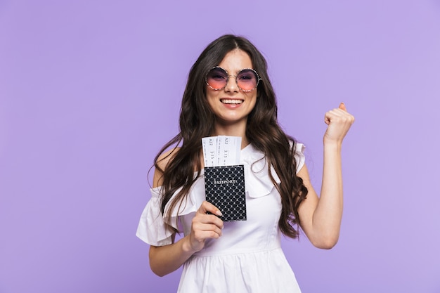 Belle jeune femme excitée portant une tenue d'été et des lunettes de soleil debout isolée sur un mur violet, montrant un passeport avec des billets d'avion