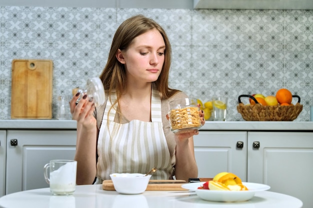 Belle jeune femme faisant une salade de fruits dans la cuisine avec pomme, orange, yaourt, flocons de maïs et graines. Alimentation saine cuisinée à la maison, beauté et santé des jeunes