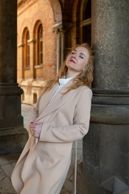 Une belle jeune femme féminine en manteau beige se tient près de la colonne d'un vieux bâtiment en fermant les yeux et en pensant à quelque chose