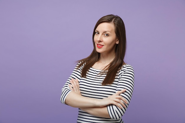 Belle jeune femme fille dans des vêtements rayés décontractés posant isolé sur fond violet violet portrait en studio. Concept de style de vie des gens. Maquette de l'espace de copie. Regardant la caméra, tenant les mains croisées.