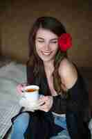 Photo belle jeune femme avec la fleur dans les cheveux. la fille est assise sur le lit et boit du thé