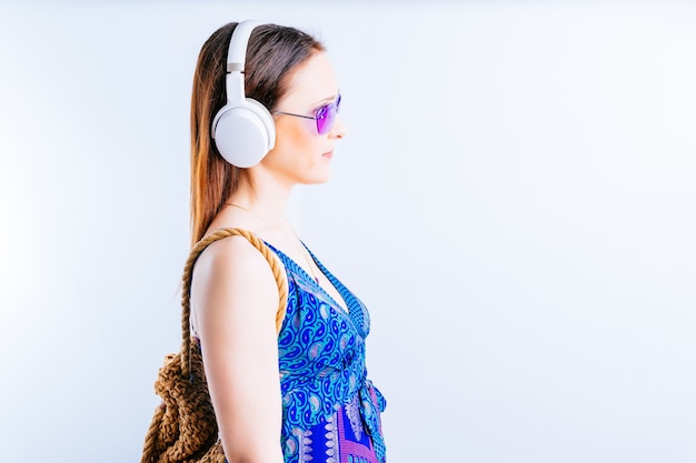 Belle jeune femme sur fond blanc vue latérale portant des lunettes d'été, écouter de la musique avec des écouteurs sans fil. concept partir en vacances