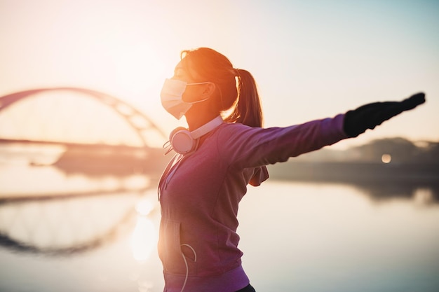 Belle jeune femme en forme en bonne forme courant et faisant du jogging seule sur la rue du pont de la ville. Elle porte un masque protecteur pour se protéger contre les virus ou les allergies. Coucher de soleil en arrière-plan.