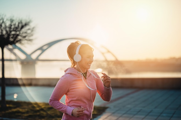 Belle jeune femme en forme en pleine forme qui court et fait du jogging seule dans la rue du pont de la ville. Elle écoute de la musique avec des écouteurs. Beau coucher de soleil en arrière-plan.